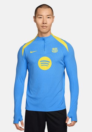 Homme portant un haut de sport bleu à manches longues avec le logo jaune Spotify et le blason du club de football de Barcelone, debout devant un fond uni.