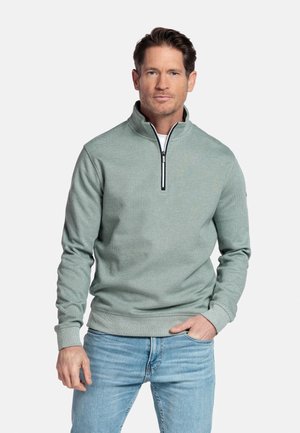 Mann trägt einen hellgrünen Pullover mit Viertel-Reißverschluss und blaue Jeans, steht mit einer Hand in der Tasche vor einem schlichten Hintergrund.