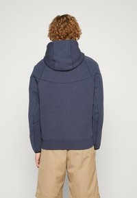 Felpa blu navy con ampio cappuccio rotondo e tessuto strutturato, con dettagli di cuciture sobrie. Abbinata a pantaloncini color sabbia, vista da dietro.