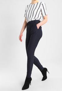 Námořnické pruhované kalhoty s vysokým pasem, spárované s bílým tričkem s krátkým rukávem, které má námořnické svislé pruhy; nošeno s černými vysokými podpatky.