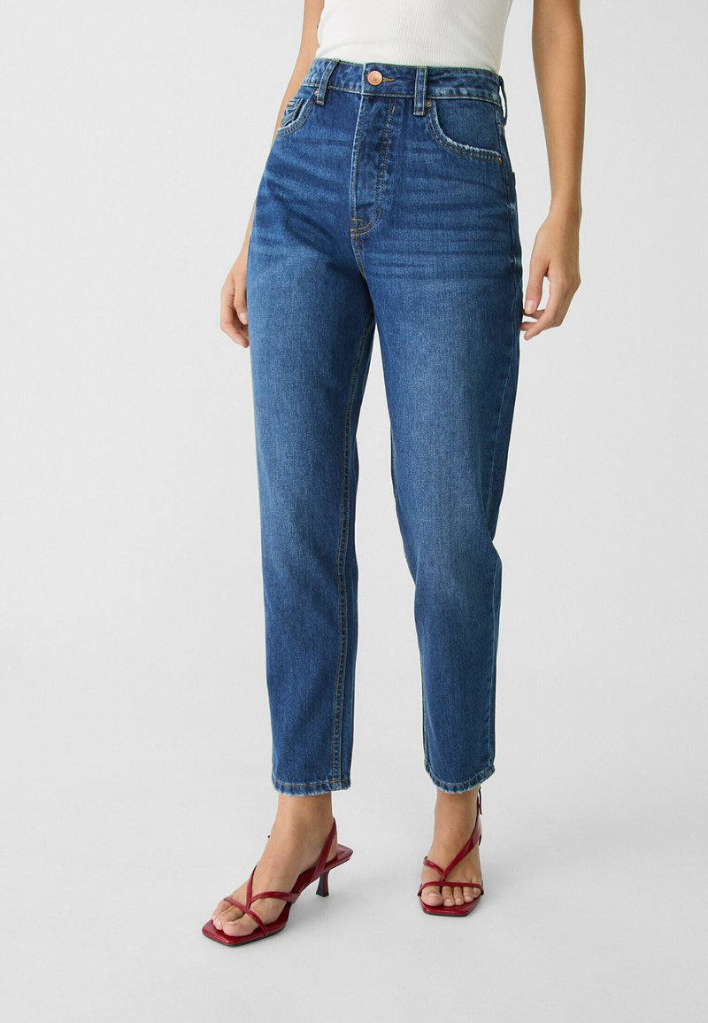 Hoch geschnittene blaue Jeans mit geradem Bein, sichtbaren Nähten, Gürtelschlaufen und Vorderentaschen. Getragen mit roten Absatzsandalen.