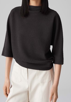 Femme portant un haut noir texturé à manches trois-quarts et un pantalon blanc ample avec poches avant sur fond gris uni.