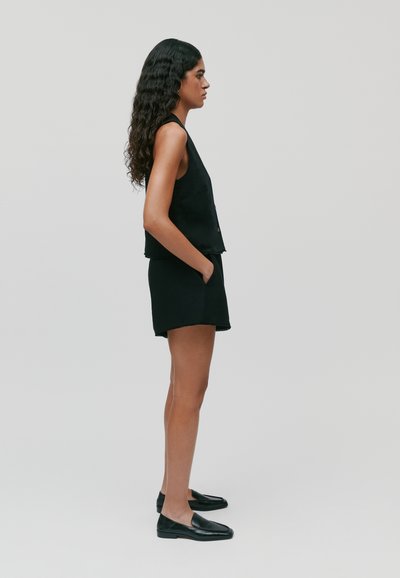 Zwarte mouwloze top gecombineerd met zwarte shorts, met een ontspannen pasvorm en rafelige randen. Model draagt zwarte loafers en staat tegen een neutrale achtergrond.