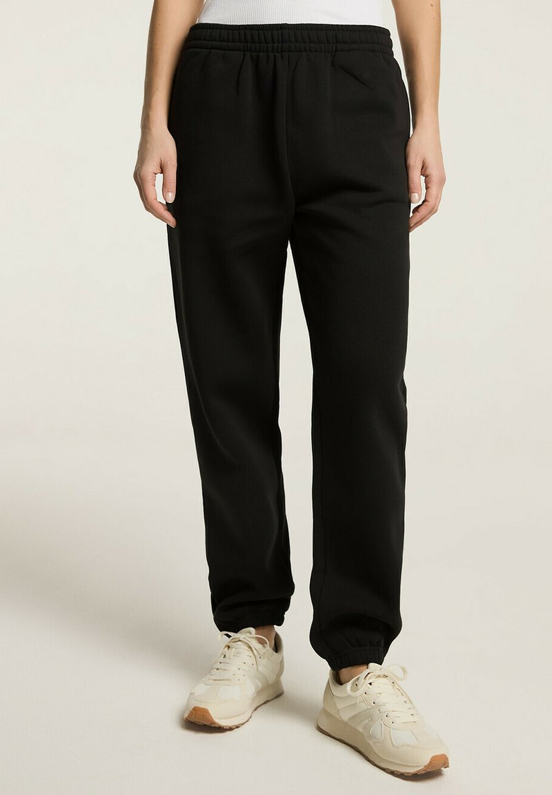 Czarne dresy z elastycznym pasem, bocznymi kieszeniami i zwężającymi się mankietami, wykonane z miękkiego materiału dla komfortu. Noszone z jasnymi sneakersami.