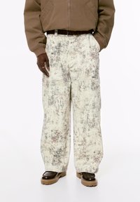 Person iført beige og brun tie-dye vide cargo bukser, brun bælte, brun jakke og mørkebrun chunky sål sko, stående med hænderne afslappet.