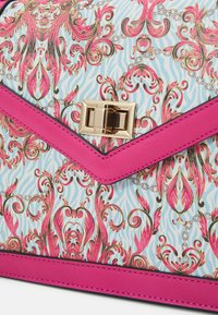 Gros plan sur le rabat d’un sac à main avec fermoir doré, bordure rose vif et motif floral baroque rose et doré complexe sur fond bleu clair.