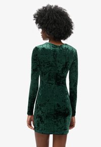 Vestido de terciopelo verde con mangas largas, diseño ajustado y textura suave. Presenta un escote redondo y un detalle de costura en la espalda.