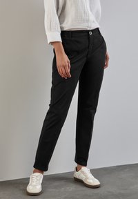 Svarta bomullsblandade byxor med slim fit, med knappstängning och bakfickor, tillsammans med vita sneakers på ett grått golv.
