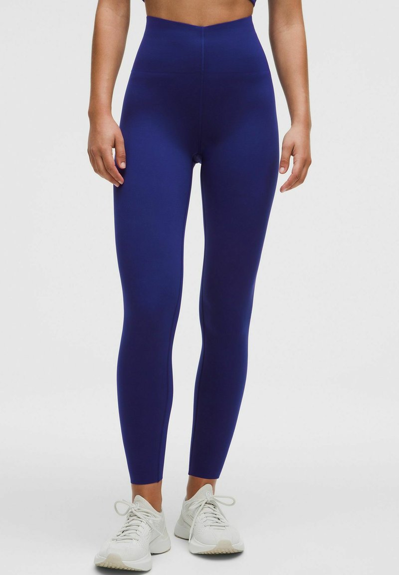 Leggings taille haute bleu marine en tissu extensible, avec une texture lisse et une coupe ajustée, associés à des chaussures de sport blanches.
