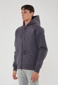 Felpa grigia con zip e cappuccio, dotata di una tasca frontale e dettaglio etichetta nera, realizzata in materiale morbido e liscio. Polsini a costine sulle maniche.
