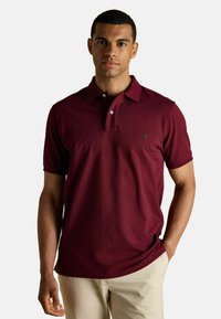 Burgundia polo särk, millel on krae, kaks nuppu ja väike roheline logo rinnal. Mugav kangas ja lühikesed varrukad. Kombineeritud heledate pükstega.