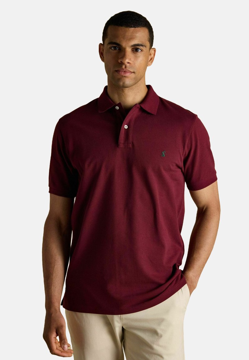 Burgundia polo särk, millel on krae, kaks nuppu ja väike roheline logo rinnal. Mugav kangas ja lühikesed varrukad. Kombineeritud heledate pükstega.