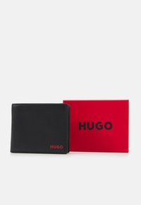 Svart läderplånbok med slät textur och röd "HUGO"-logotyp på framsidan, placerad bredvid en röd låda med samma logotyp.