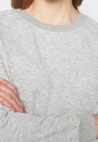 Sudadera gris claro con superficie texturizada, cuello redondo acanalado y puños. Presenta costuras visibles y un ajuste holgado.