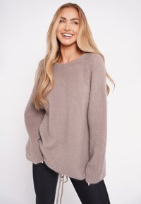 Jasno-beżowy, oversize sweter o miękkiej, puszystej fakturze, z okrągłym dekoltem i sznurkiem przy dolnym brzegu, zestawiony z czarnymi legginsami.