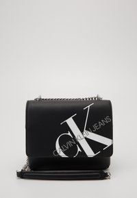 Černá kožená crossbody taška se stříbrným řetízkem a velkým bílým logem CK a textem "Calvin Klein Jeans" na přední části.