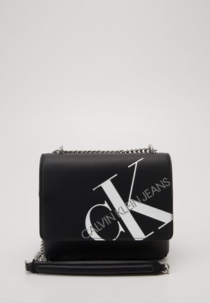 Sac en cuir noir porté croisé avec chaîne en argent et grand logo CK blanc ainsi que le texte "Calvin Klein Jeans" sur le rabat avant.