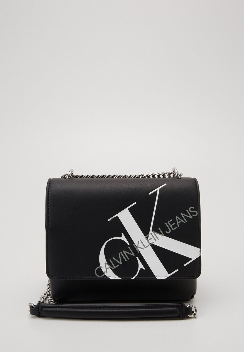 Černá kožená crossbody taška se stříbrným řetízkem a velkým bílým logem CK a textem "Calvin Klein Jeans" na přední části.