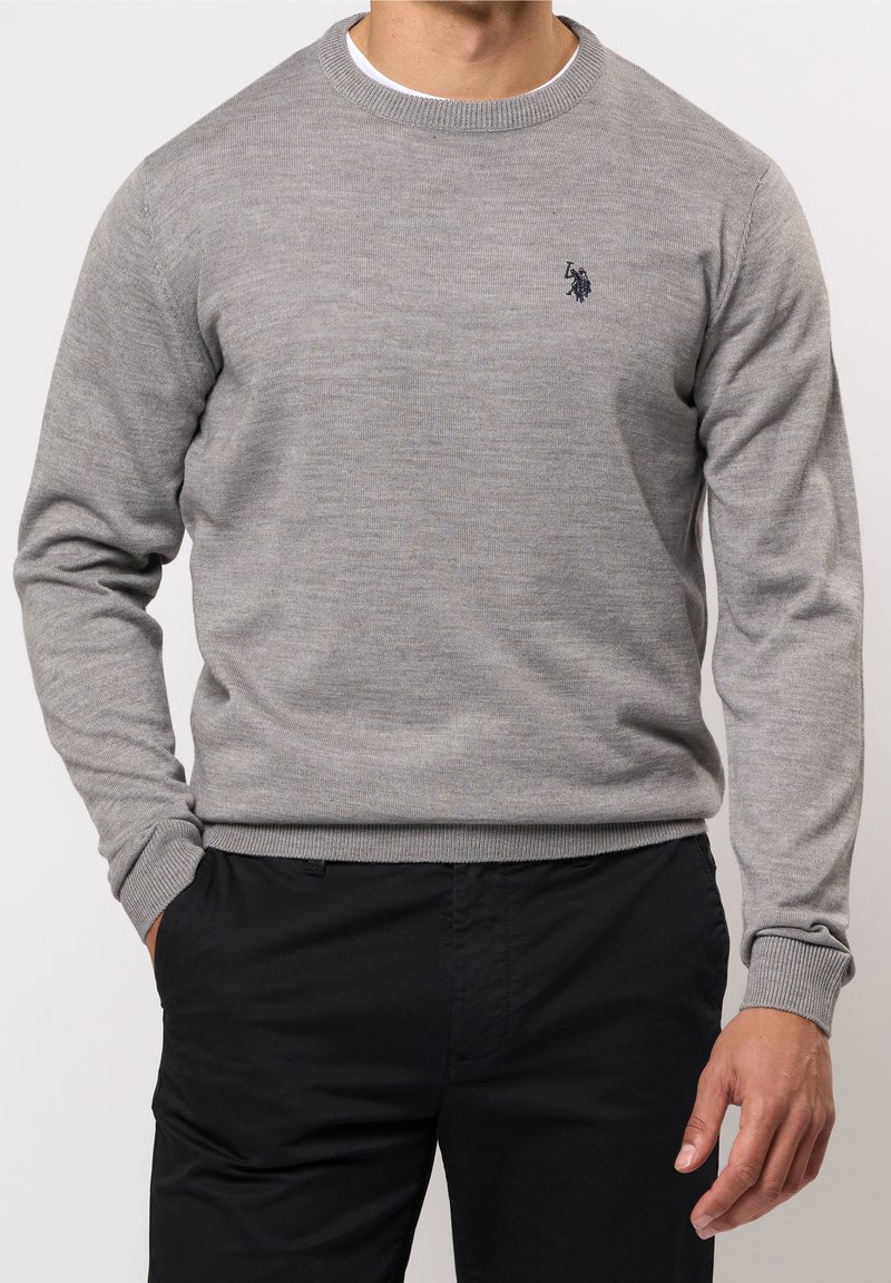 Pull gris en maille avec un col rond et des poignets côtelés, présentant un petit logo sombre sur la poitrine. Porté avec un pantalon noir.