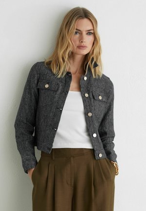 Femeie blondă purtând o jachetă texturată neagră cu nasturi, top alb cu dungi și pantaloni maro cu talie înaltă, cu mâinile în buzunare, în fața unui perete alb.