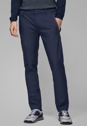 Mann trägt marineblaue schlanke Hose, marineblauen Pullover und schwarz-weiße Sneakers, steht mit einer Hand in der Tasche vor grauem Hintergrund.