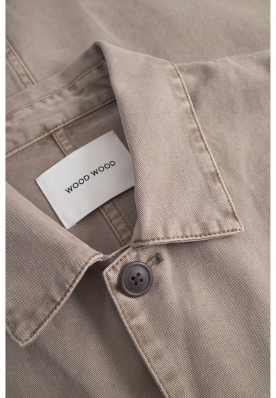 Beige gekraagde shirt van lichtgewicht stof, met een grijze knop en een wit label met "WOOD WOOD" in zwarte tekst.
