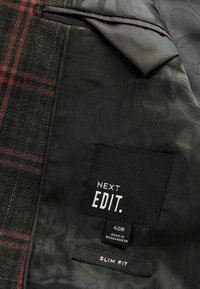 Czarna tkanina z szarym wzorem kwiatowym, z tartanowym sekcją w czerwone i białe linie. Metki informują o "NEXT EDIT", rozmiar 40R oraz "SLIM FIT".