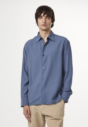 Long-sleeve shirt in light blue fabric with a textured finish, buttoned down front, and standard collar. Styled with khaki pants.