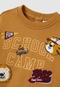 Sudadera de algodón marrón con un cuello acanalado, que presenta parches bordados coloridos de un león, un tigre y el texto "CAMPAMENTO ESCOLAR" y "Mejor equipo."