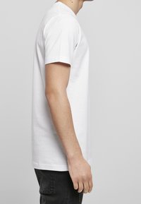 Boční pohled na osobu v obyčejném bílém tričku s krátkým rukávem a černých kalhotách na šedém pozadí.