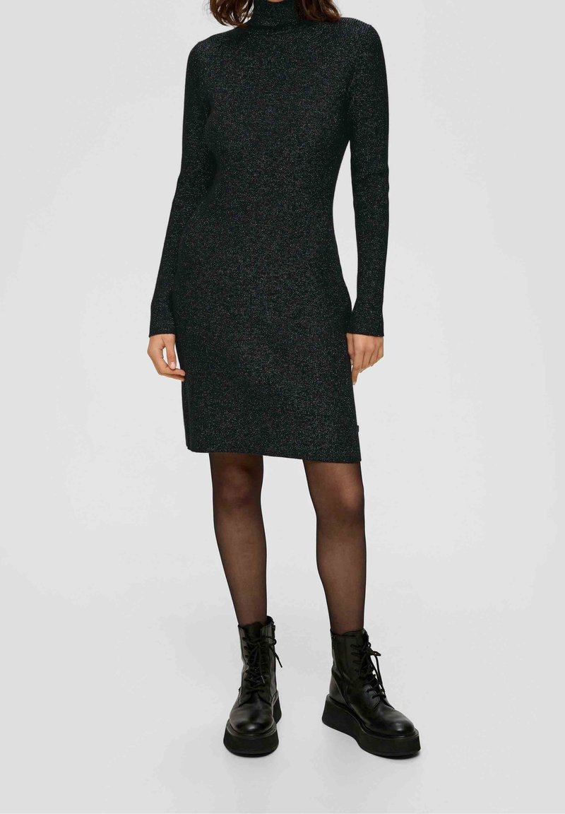 Femme portant une robe noire à col roulé à manches longues, des collants noirs transparents et des bottes à plateforme noires à lacets, debout sur un fond clair.
