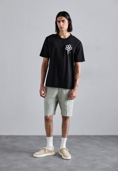 Schwarzes Baumwoll-T-Shirt mit weißem Blumenmuster, kombiniert mit hellgrünen Shorts. Die Schuhe sind beigere Sneaker mit weißen Gummisohlen und weißen Socken.