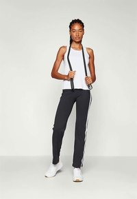 Balta tops, melni sporta bikses ar baltu maliņu un baltas sporta apavus. Modelis tur melnas siksnas uz neitrāla fona.