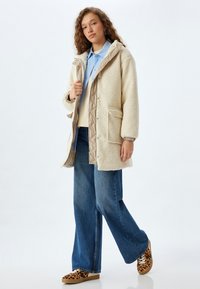 Krämlik shearlingjacka med en beige quiltad foder, oversize passform, framfickor, bärs över en ljusblå skjorta och lösa blå jeans med leopardmönstrade sneakers.