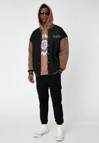 Chaqueta de varsity negra y marrón con puños a rayas sobre una sudadera marrón con capucha. Pantalones cargo oscuros y zapatillas altas blancas completan el look.