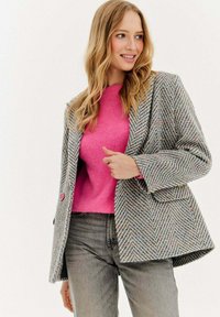 Blondýnka oblečená v růžovém svetru, šedém vzorovaném blejzru s barevnými tečkami a vybledlých šedých džínách, usmívající se a koukající na stranu.