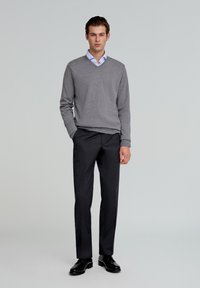Šedý svetr s výstřihem do V přes světle modrou košili, doplněný o tmavé slim kalhoty a černé boty. Hladká textura a řemeslný design.