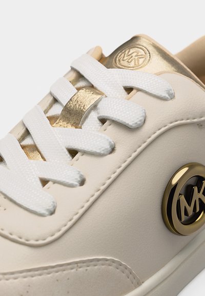 Κοντινή λήψη μπεζ αθλητικού παπουτσιού με λευκά κορδόνια, χρυσά στόμια και μεταλλικό έμβλημα λογότυπου στο πλάι.