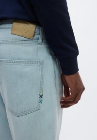 Τζιν σε ανοιχτό μπλε χρώμα με ταμπά δερμάτινο έμπλαστρο και πολύχρωμες ραφές, με πίσω τσέπη και ίσια γραμμή παντελονιού.