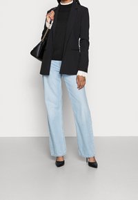 Blazer noir sur mesure porté sur un pull noir, associé à un jean bleu clair à jambes larges et des escarpins noirs à bouts pointus. Porte un sac à main noir.