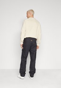 Mann mit kurzen blonden Haaren, der einen beigen Sweatshirt, schwarze Cargo-Hosen und weiße Turnschuhe trägt, steht vor einer schlichten weißen Wand.