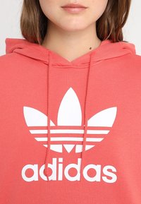 Korálově růžová mikina Adidas má velké bílé logo trefoil a tři vodorovné pruhy, s kapucí na šňůrku a měkkou texturou materiálu.