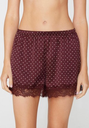 Frau trägt bordeauxrote Shorts mit Polka-Dots und Spitzenbesatz am Saum, Hände entspannt seitlich, sichtbarer Bauch und obere Oberschenkel.