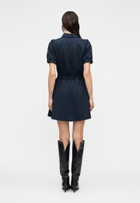 Γυναίκα με βρεγμένα μαλλιά, που φοράει ένα ναυτικό μπλε φόρεμα με κοντά μανίκια και μαύρες μπότες με τακούνι μέχρι το γόνατο, στέκεται γυρισμένη προς τα πίσω σε ένα απλό φόντο.