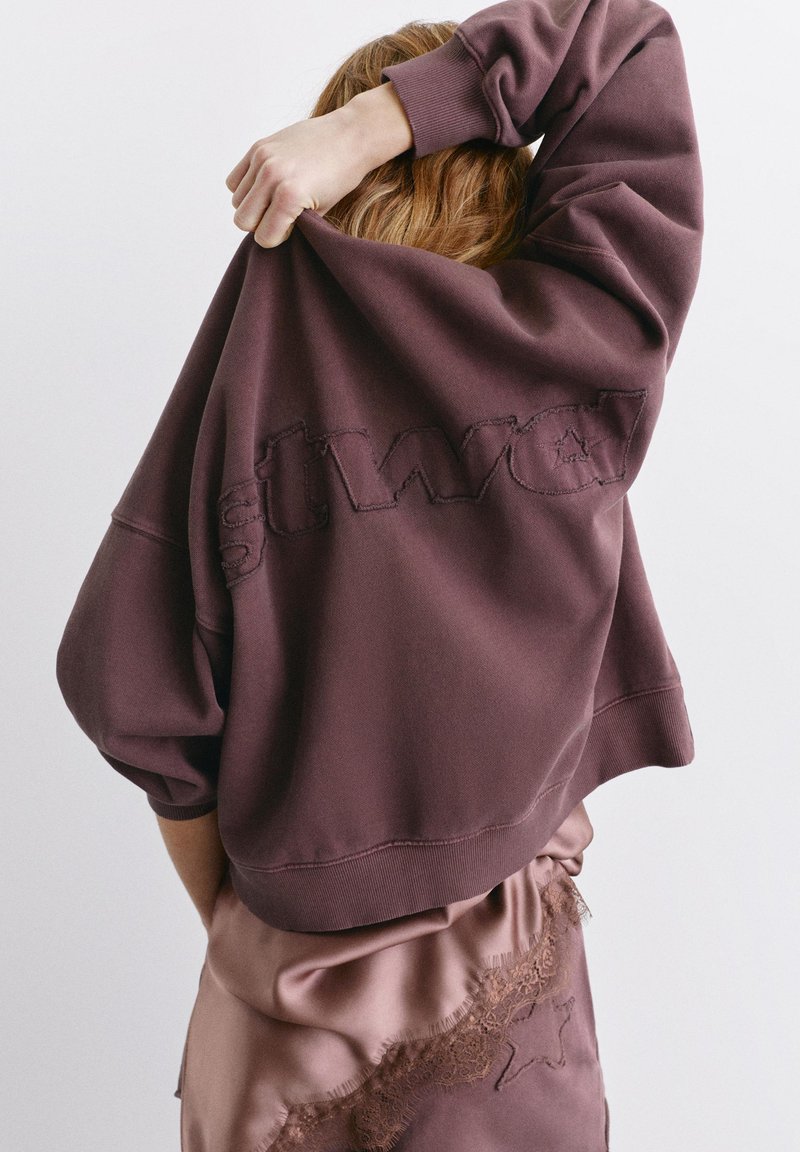 Personne en train d’enfiler un sweat-shirt violet foncé par-dessus la tête, portant une jupe en satin avec des détails en dentelle et une broderie d’étoiles.