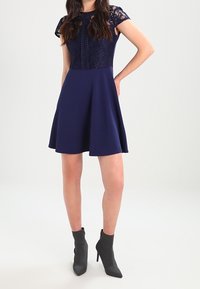 Abito blu navy con corpino in pizzo, maniche a sbuffo corte e vita aderente; gonna svasata. Indossato con stivaletti grigi alla caviglia, che evidenziano una silhouette elegante.