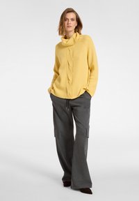 Gelber Strickpullover mit einem Kragen und Zopfmuster, kombiniert mit grauen weit geschnittenen Hosen mit Seitentaschen und einem Kordelzug in der Taille.