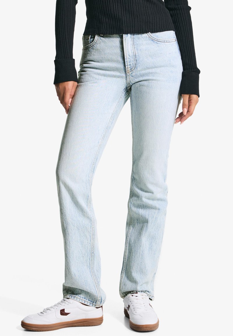 Personne portant un jean droit bleu clair, un pull noir côtelé et des baskets blanches aux semelles marron, debout devant un fond uni.
