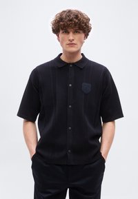 Νεαρός άνδρας με σγουρά μαλλιά στέκεται προς τα εμπρός, φορώντας ένα μαύρο πουκάμισο με κοντά μανίκια, κουμπωτό και με γιακά, με λεπτές κάθετες ρίγες.