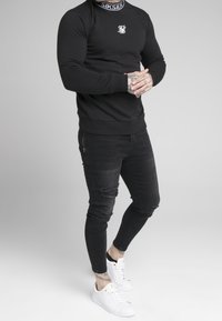 Man i svart långärmad skjorta, trasiga svarta skinny jeans, vita sneakers och tatuerade händer som hålls ihop framför mot en neutral bakgrund.